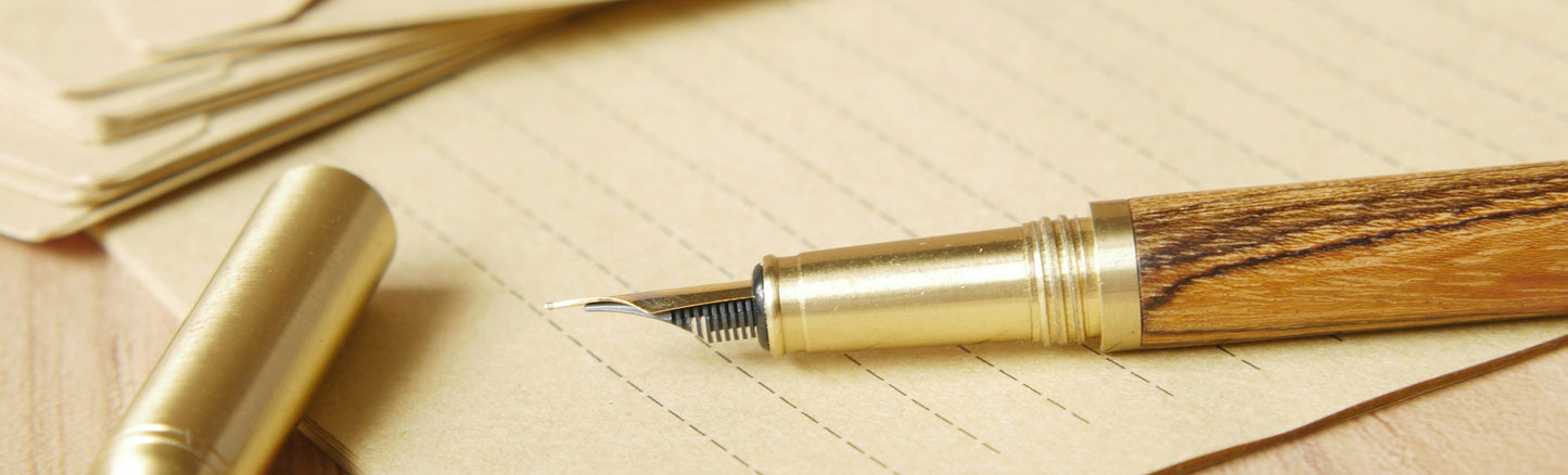 l'image montre un beau stylo plume en bois et métal doré posé sur un papier de qualité sur une table en bois. L'ambience autour de ce bel intstrument d'écriture se veut chaleureuse invitant à l'écriture manuelle, exprimant le plaisir d'écrire à l'encre. 